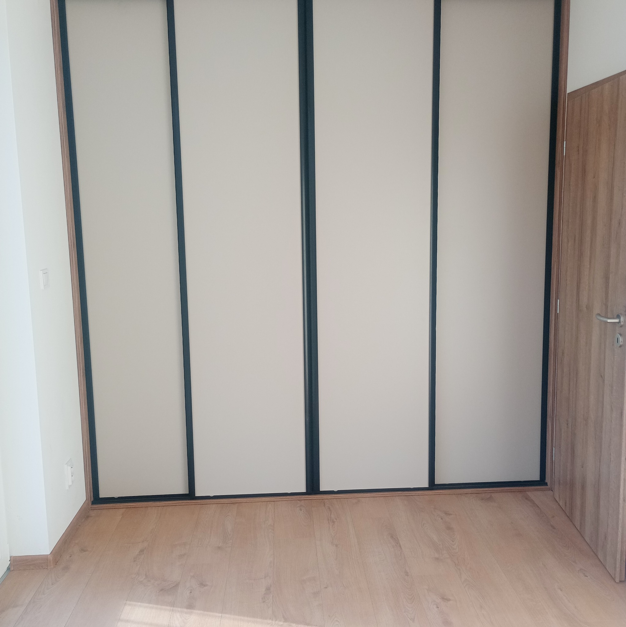 Czterodrzwiowa szafa wnękowa z przesuwnymi, beżowymi frontami i czarnymi obramowaniami, na tle drewnianej podłogi i fragmentu ściany z drzwiami.