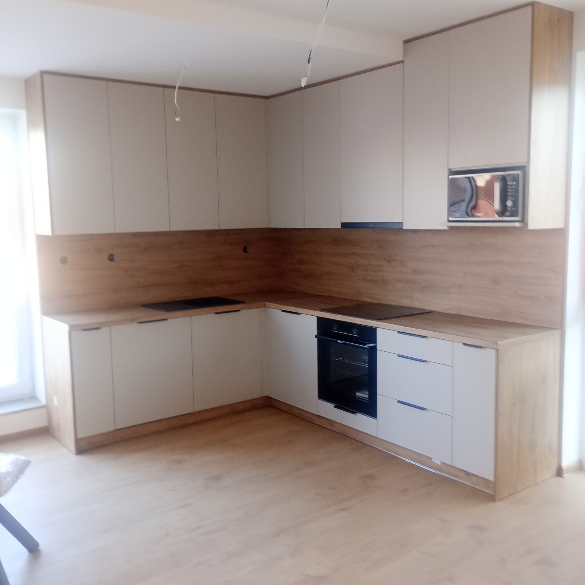 Jasna kuchnia narożna z drewnianym blatem i białymi frontami szafek. Widoczny piekarnik, płyta indukcyjna i kuchenka mikrofalowa. Minimalistyczny design.