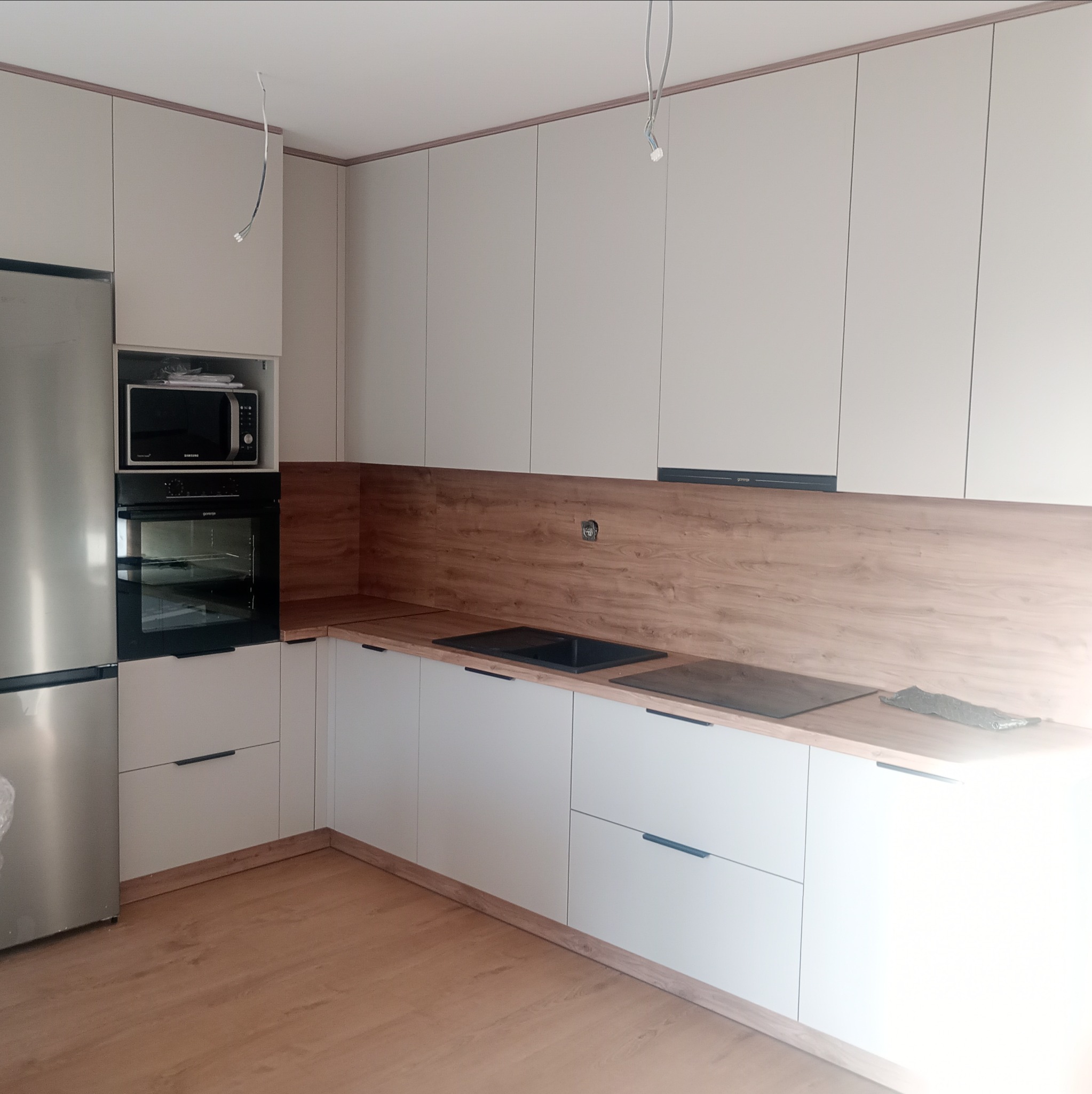 Jasna kuchnia na wymiar z drewnianym blatem i sprzętami AGD. Widoczne wiszące kable oświetleniowe. Minimalistyczny design z długimi, czarnymi uchwytami.