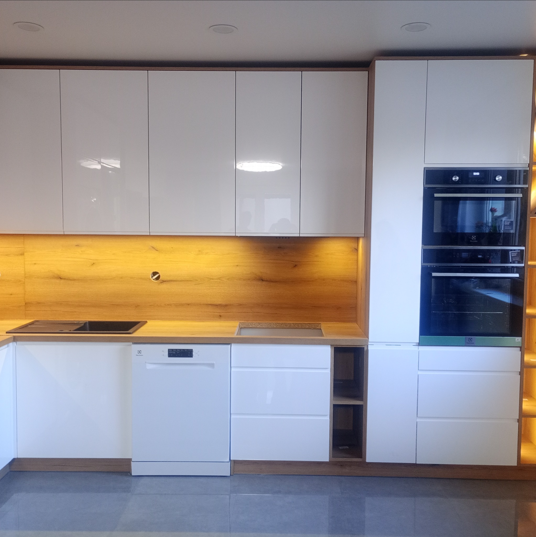 Nowoczesna kuchnia z białymi, lakierowanymi frontami i drewnianym panelem ściennym z oświetleniem LED. Widoczne wbudowane sprzęty AGD.