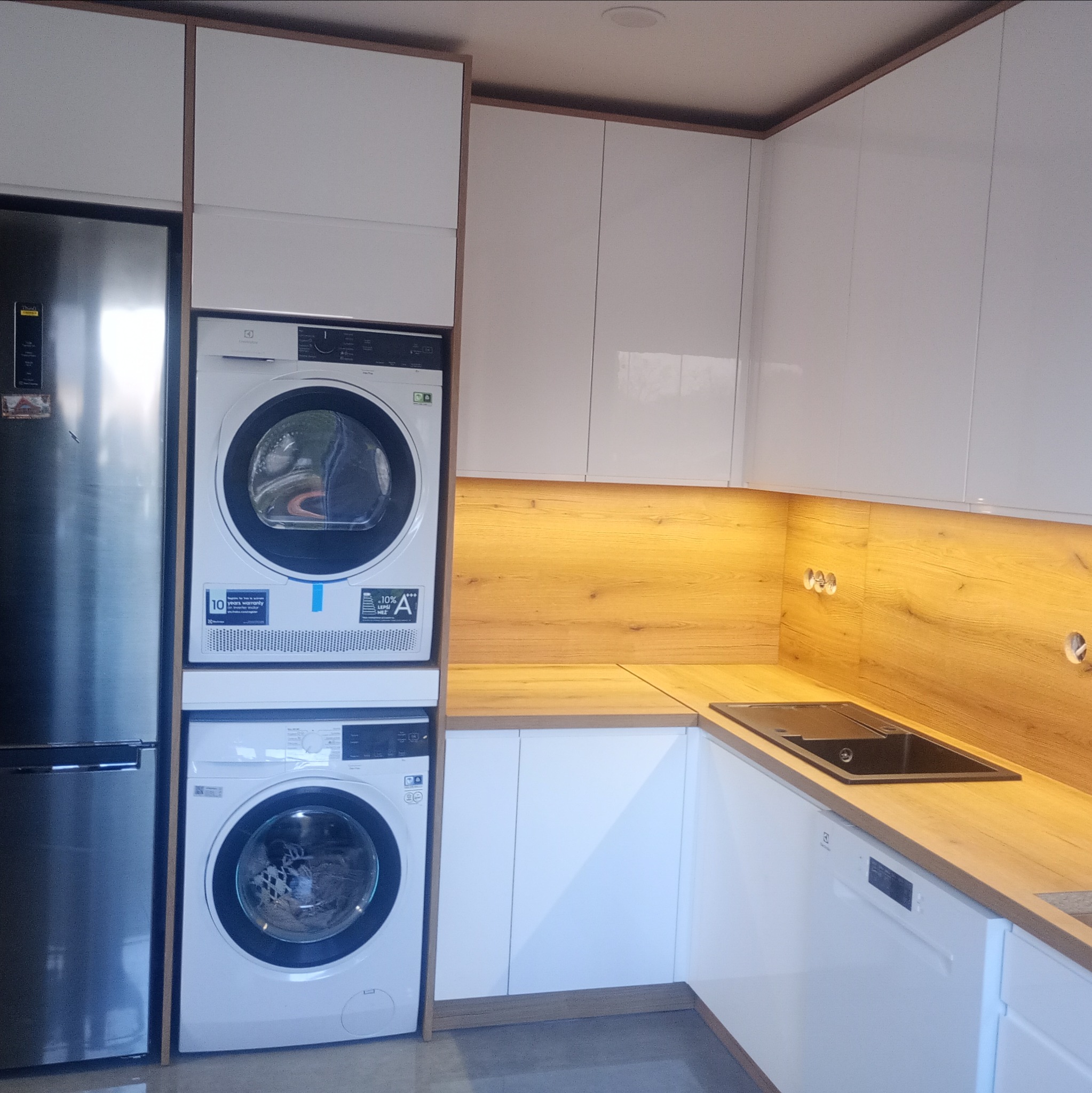 Biała zabudowa kuchenna z drewnianym blatem, zintegrowana z pralką i suszarką Electrolux, obok lodówka. Ujęcie z perspektywy wnętrza kuchni, widoczny zlew.