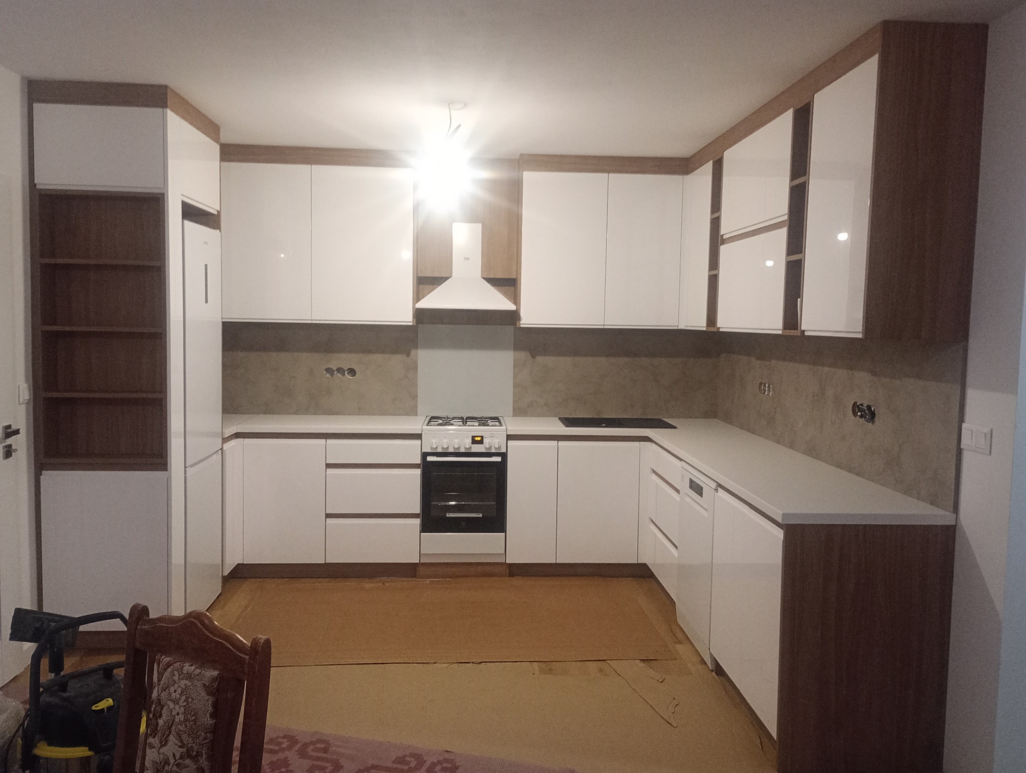 Nowoczesna kuchnia z białymi frontami i drewnianymi akcentami. Widoczny sprzęt AGD, jasne blaty i oświetlenie sufitowe. Na podłodze zabezpieczenie kartonem.