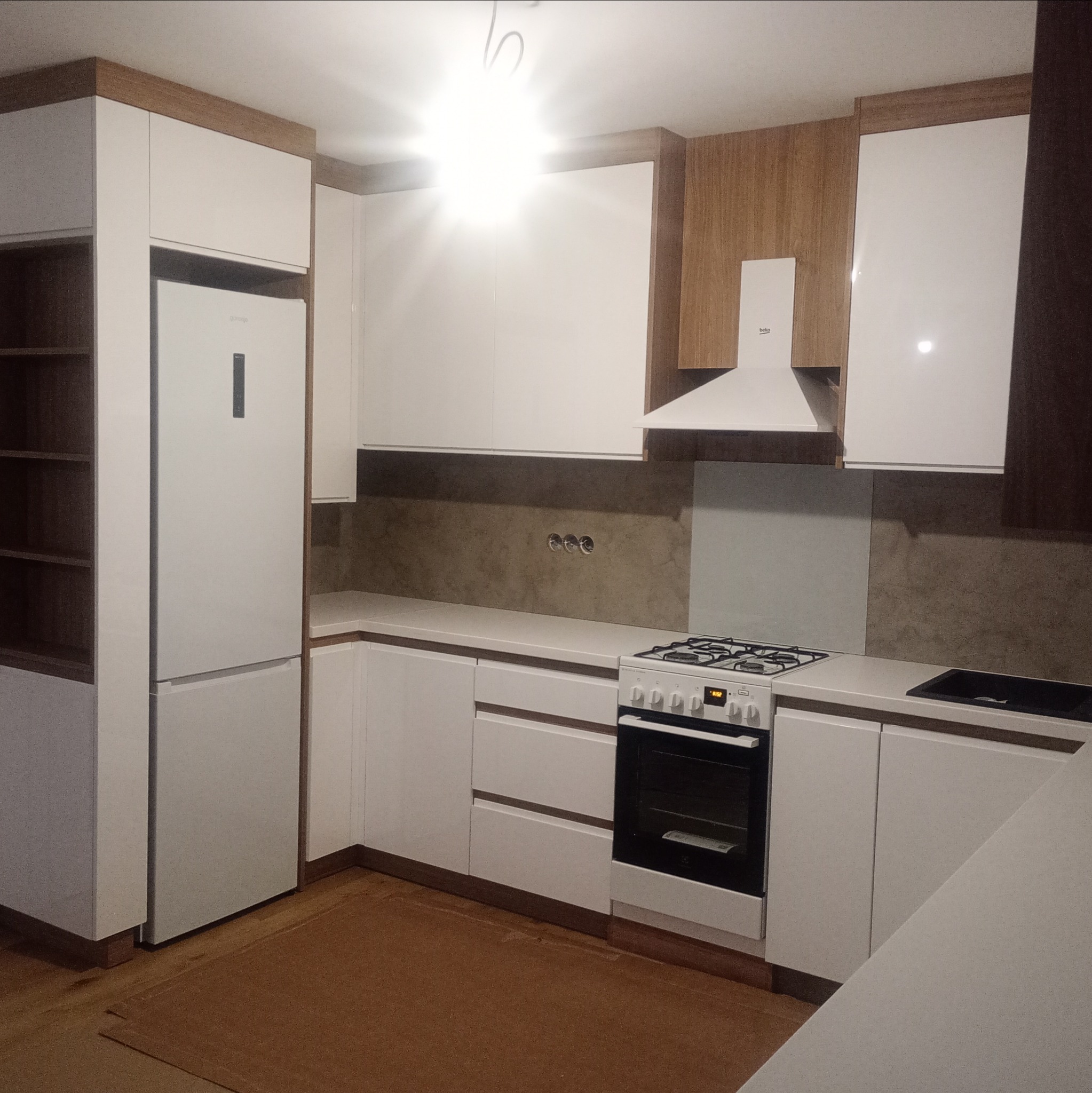 Nowoczesna, jasna kuchnia z białymi frontami i drewnianymi akcentami. Widoczna zabudowa z lodówką, kuchenką i okapem. Uwagę zwraca brązowy dywan na podłodze.