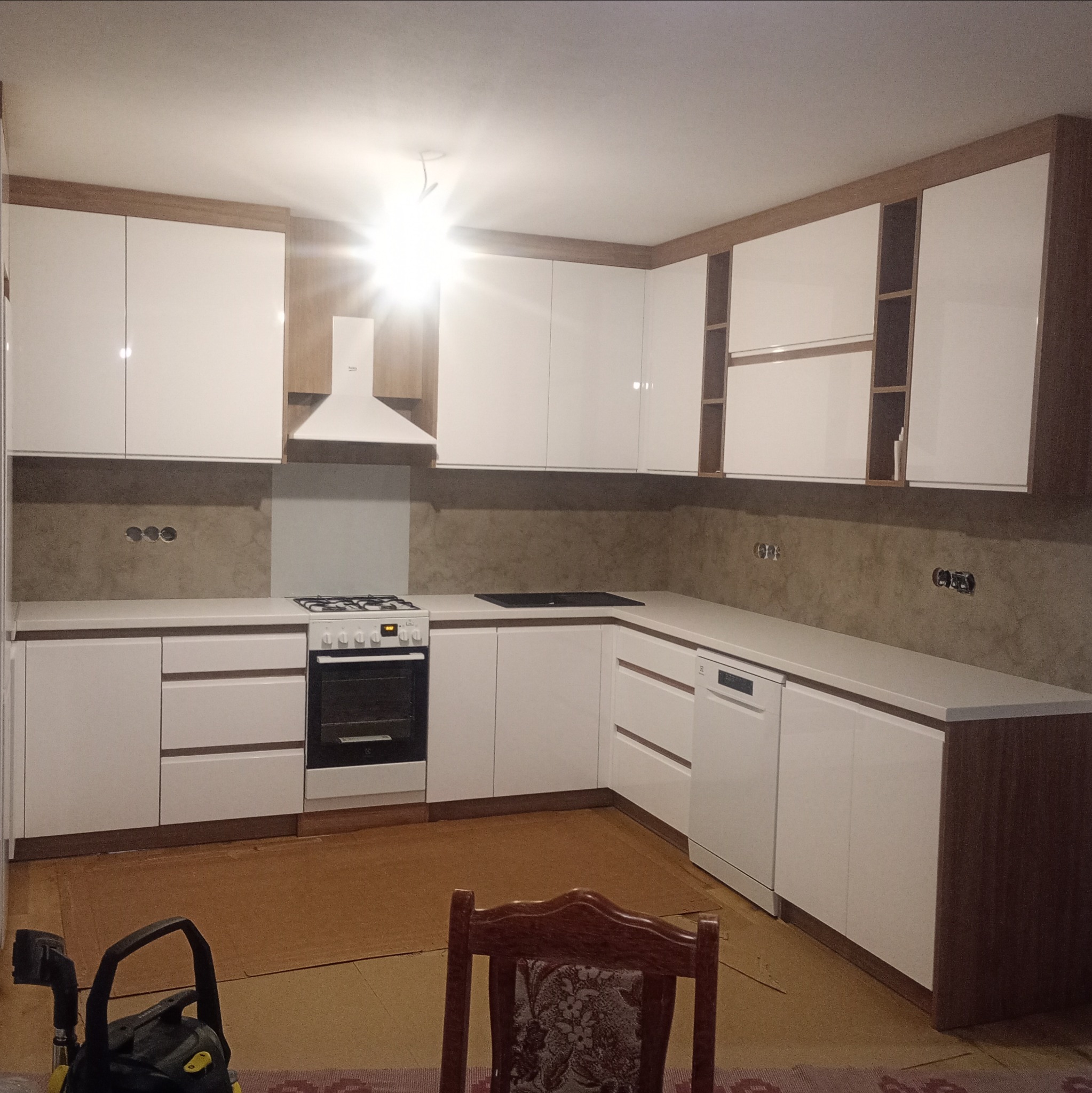 Nowoczesna kuchnia na wymiar z białymi frontami i drewnianymi akcentami. Widoczny sprzęt AGD: piekarnik, płyta grzewcza, okap i zmywarka. Oświetlenie sufitowe.