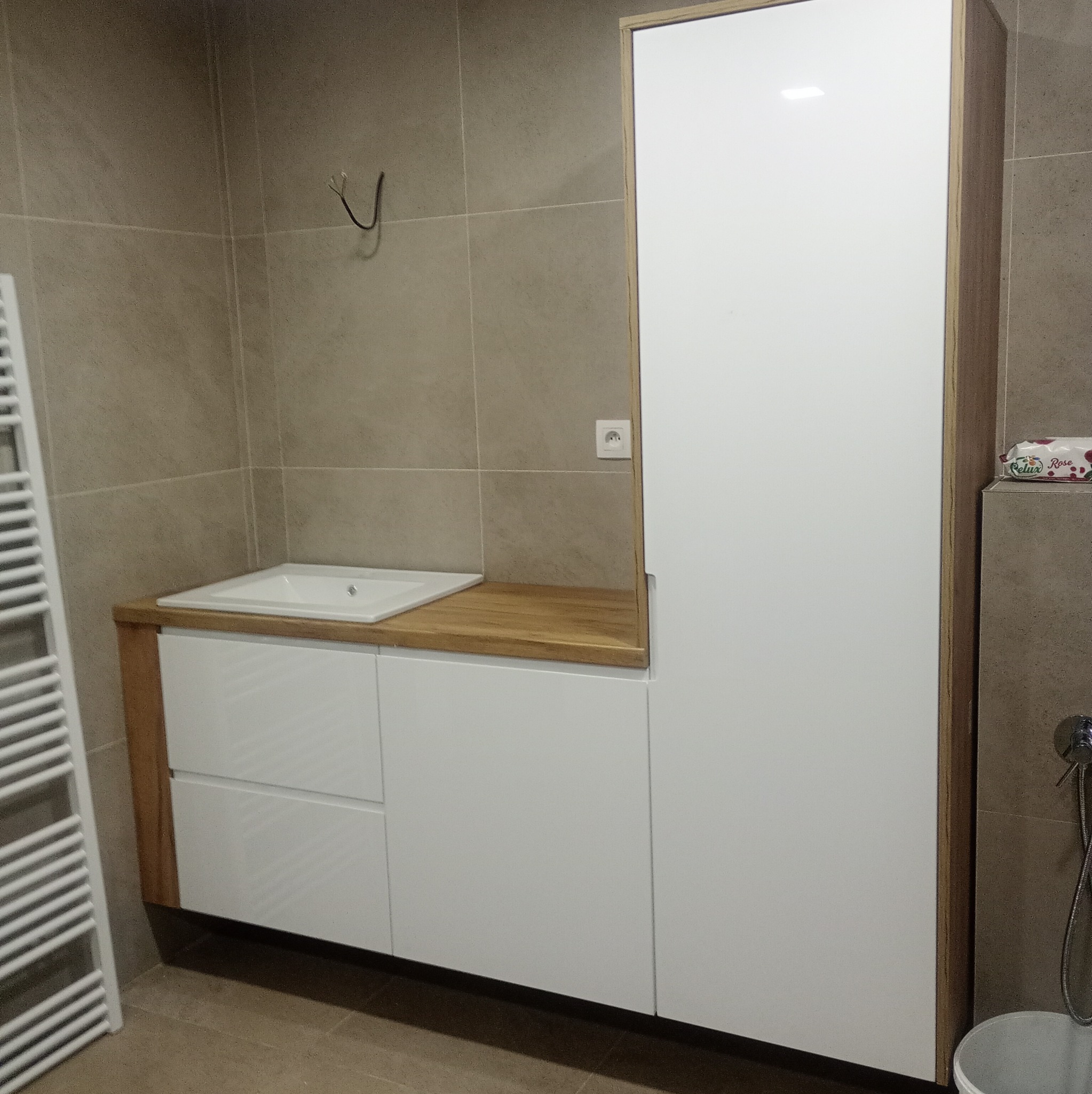 Nowoczesna łazienka z szafką pod umywalką z drewnianym blatem i wysoką szafką. Minimalistyczny design, beżowe płytki, grzejnik drabinkowy.