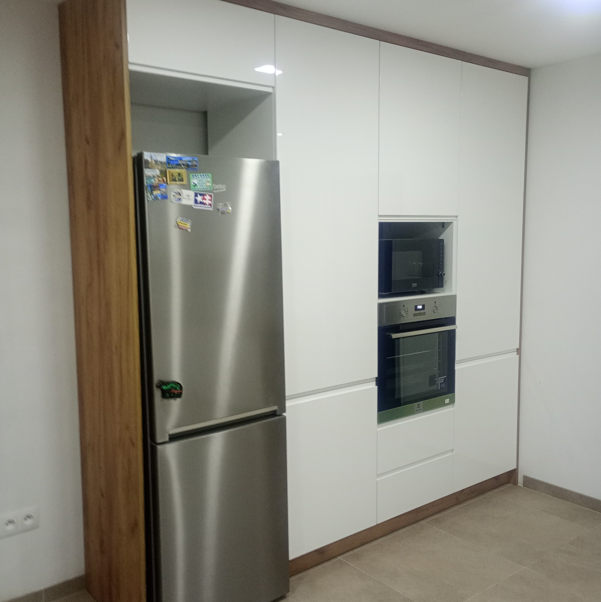 Biała zabudowa kuchenna z drewnianym akcentem, wbudowanym piekarnikiem i mikrofalówką. Obok lodówka z magnesami. Minimalistyczny design kuchni.