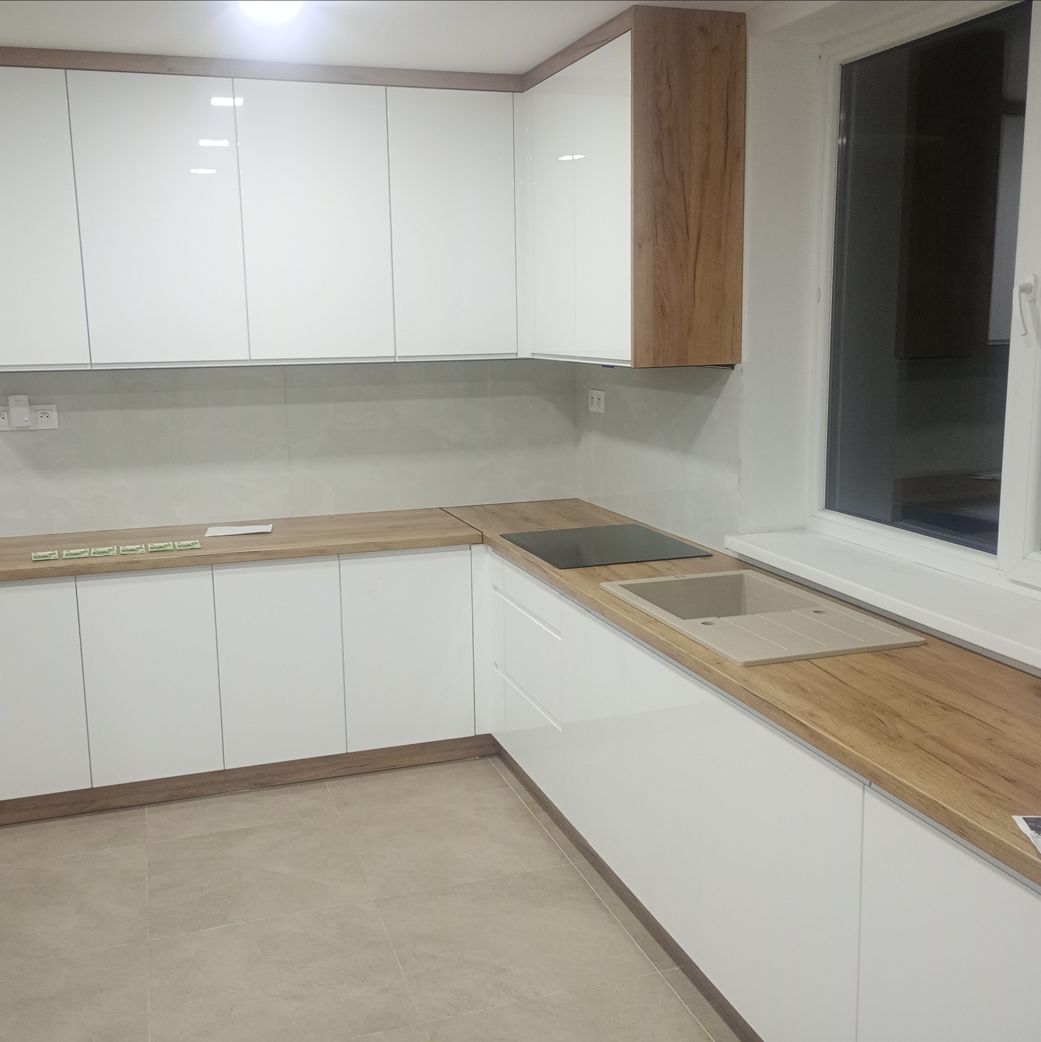 Jasna kuchnia z białymi szafkami i drewnianym blatem, zlewem i płytą. Widoczne okno i beżowe płytki podłogowe, minimalistyczny design.