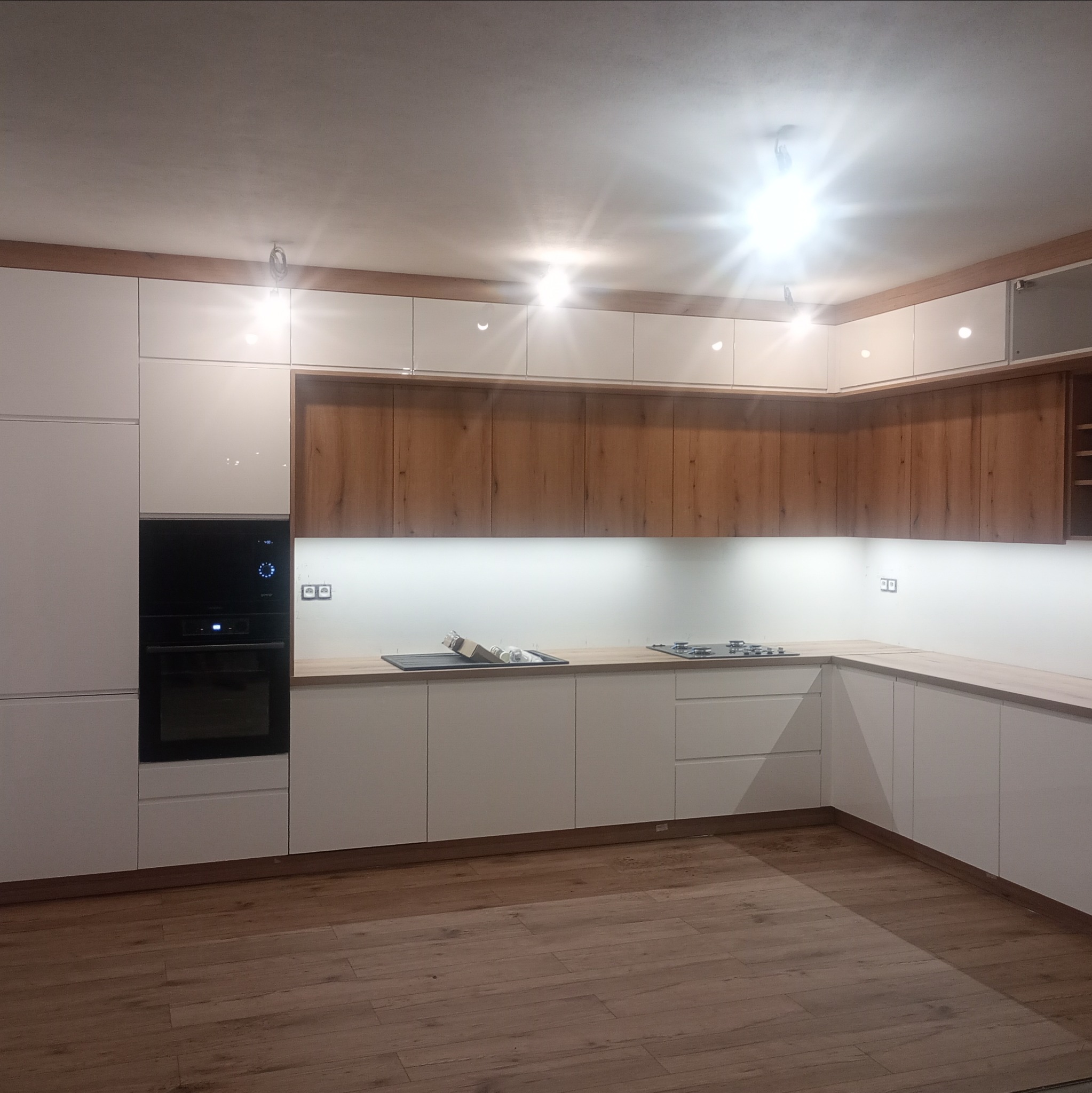 Nowoczesna, jasna kuchnia z drewnianymi akcentami w Barwałdzie Górnym. Zabudowa z piekarnikiem, płyta grzewcza i drewnopodobna podłoga. Górne szafki podświetlone.