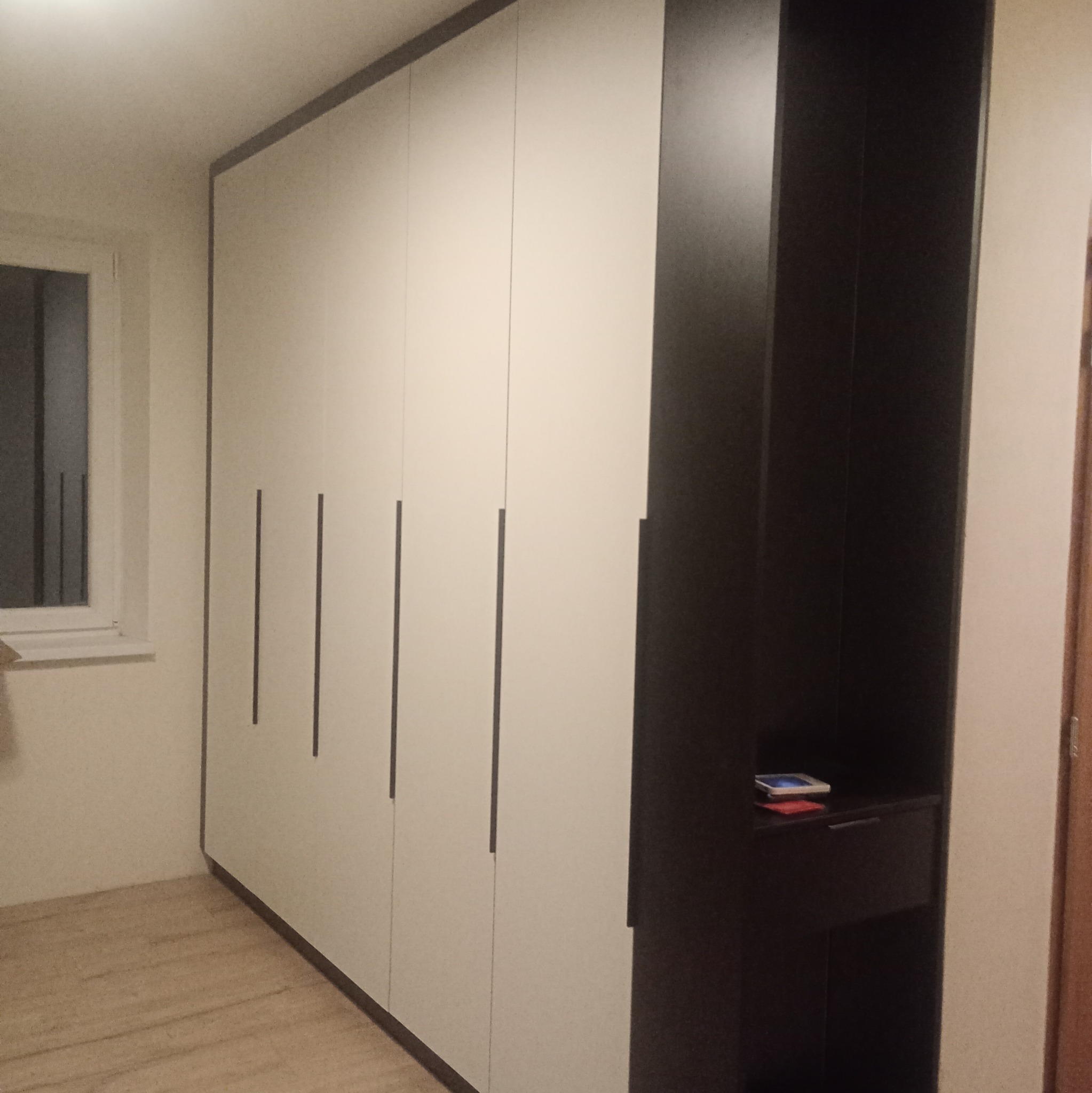 Biała szafa wnękowa z czarnymi akcentami i pionowymi uchwytami, widoczna z boku, z małą półką z przegródkami po prawej stronie. Minimalistyczny design.