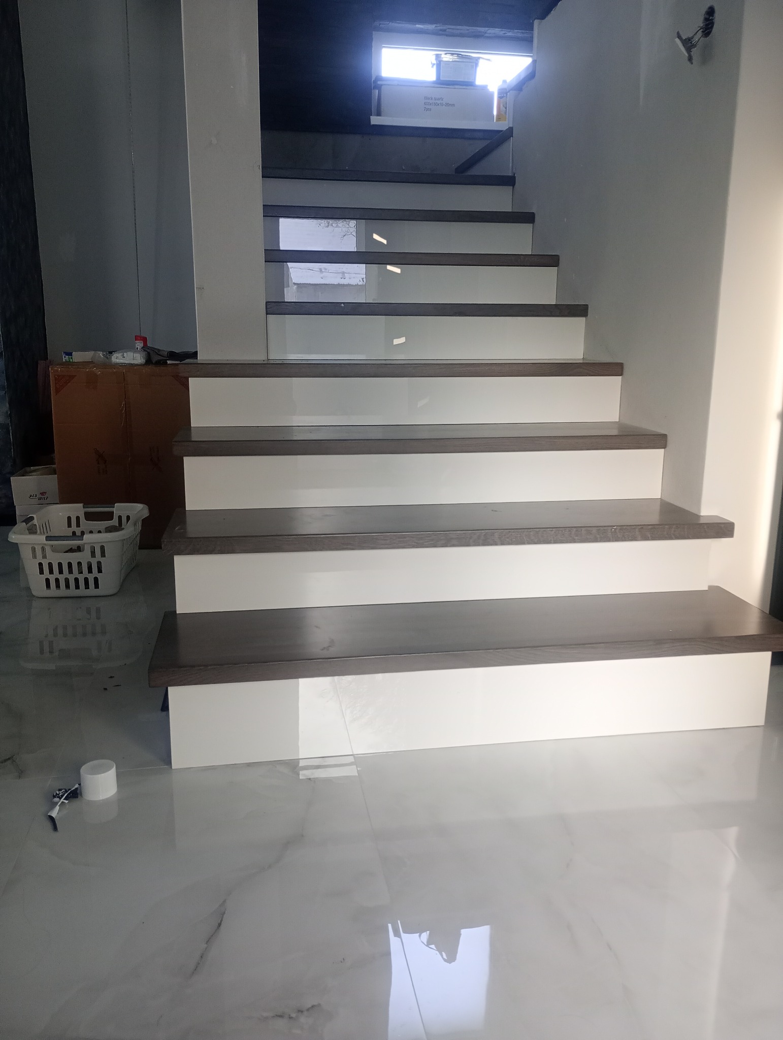 Nowoczesne schody z drewnianymi stopniami i białymi podstopnicami, widok z dołu, marmurowa podłoga odbija światło. W tle kartony i akcesoria montażowe.