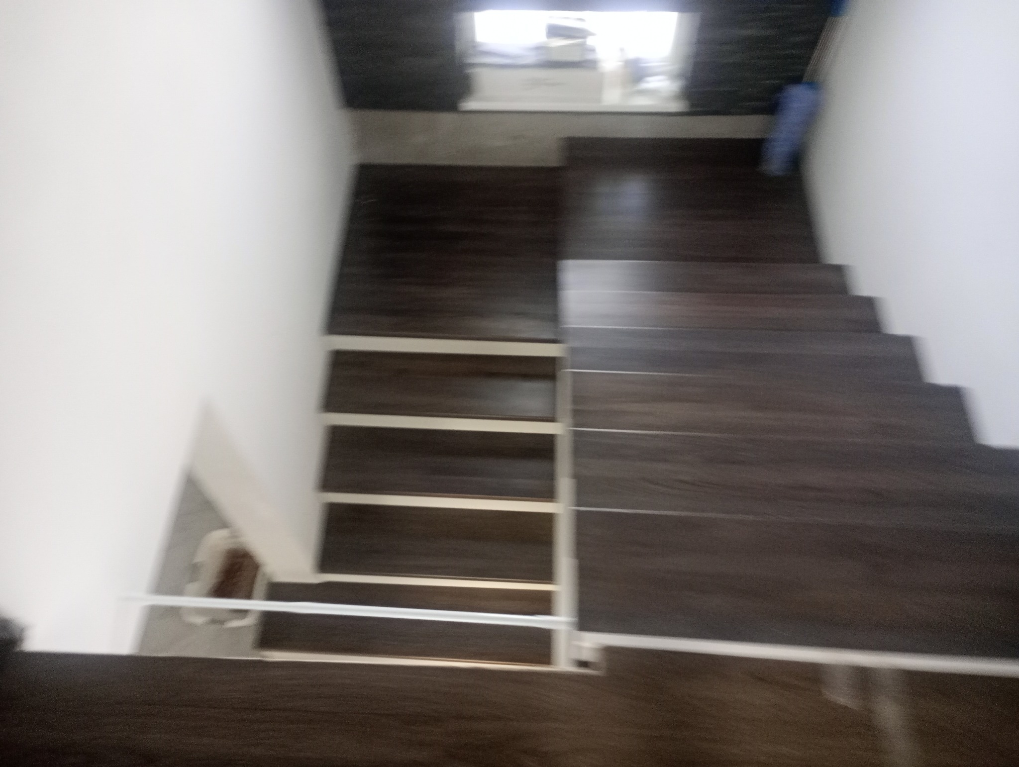 Widok z góry na drewniane schody z białymi barierkami, prowadzące do góry. Ciemne drewno kontrastuje z białymi ścianami. Na górze widoczne okno.