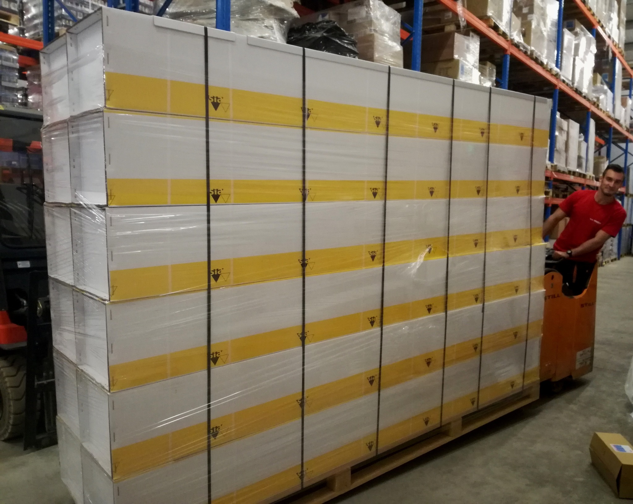 Paleta z zapakowanymi kartonami z żółtym pasem i logo w magazynie, obsługiwana przez operatora wózka widłowego. Widoczne regały magazynowe w tle.
