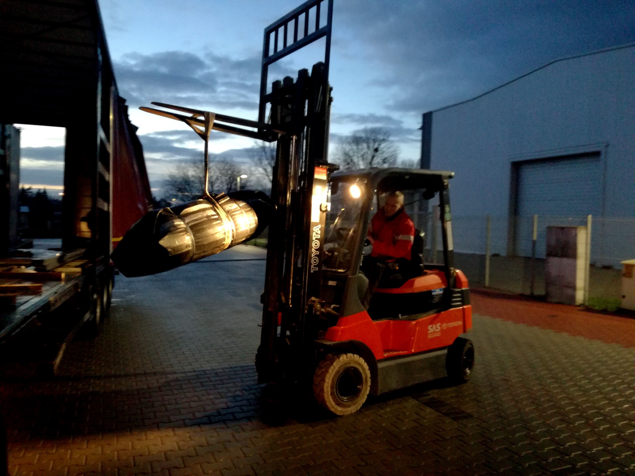 Czerwony wózek widłowy Toyota SAS unosi owinięty ładunek na tle magazynu i nieba o zmierzchu. Operator w czerwonej kurtce w kabinie.