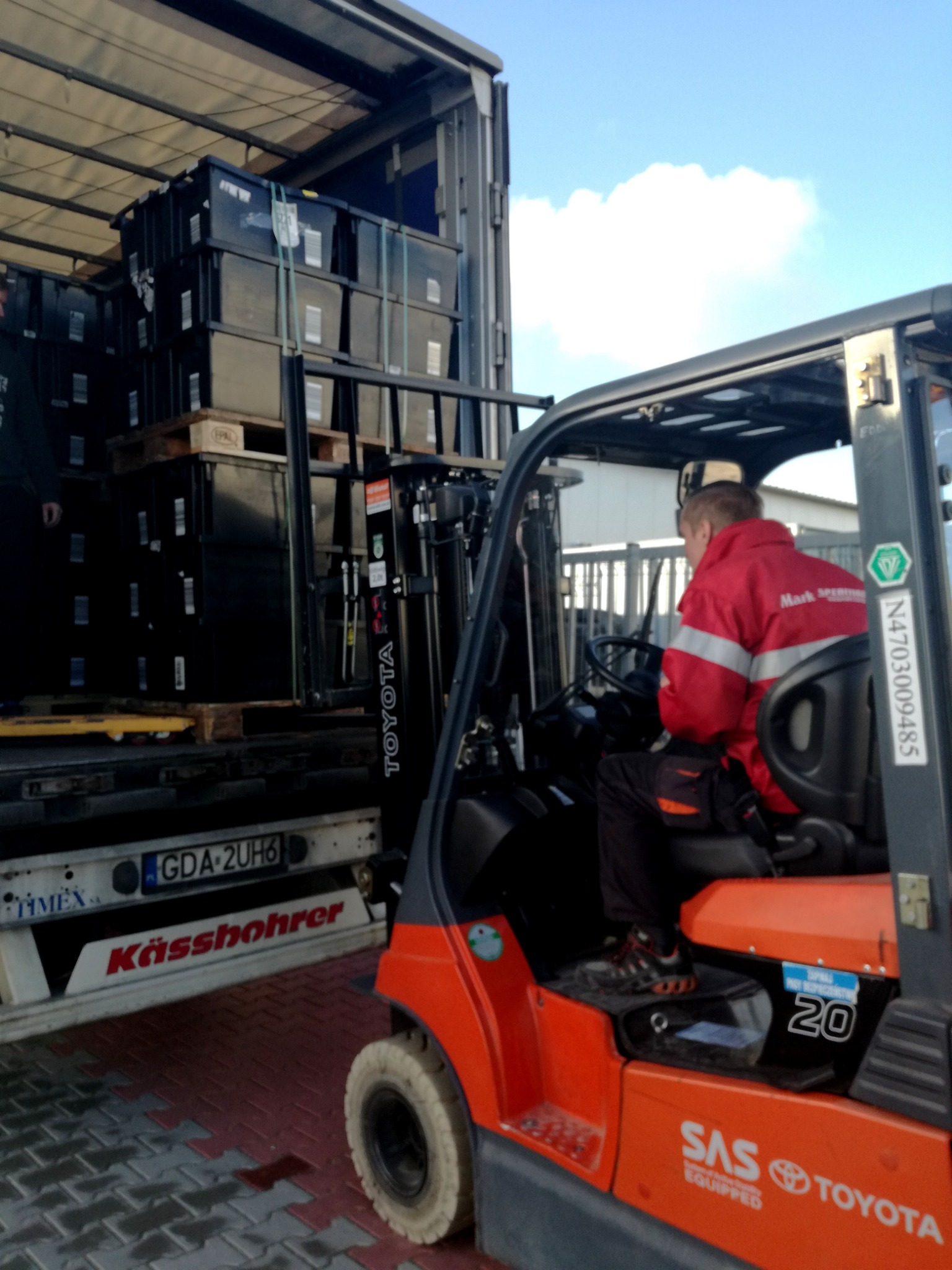 Wózek widłowy Toyota ładuje czarne pojemniki na paletach do naczepy ciężarówki. Widoczny operator w czerwonej kurtce i tablica rejestracyjna GDA-2UH6.