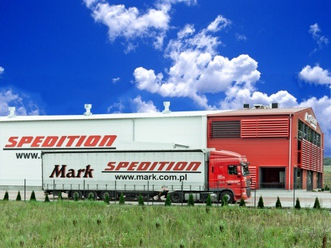 Czerwona ciężarówka z naczepą Mark Spedition parkuje przed magazynem z logo firmy na tle błękitnego nieba z chmurami i zieleni.