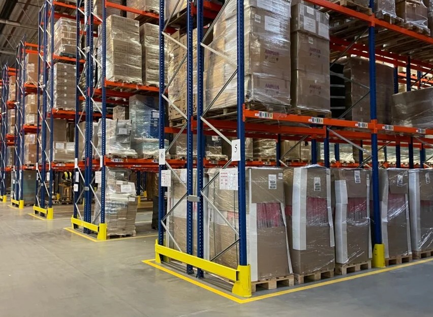 Regały magazynowe z zapakowanymi kartonami, zabezpieczonymi folią. Widoczne numery regałów i żółte bariery ochronne na podłodze.