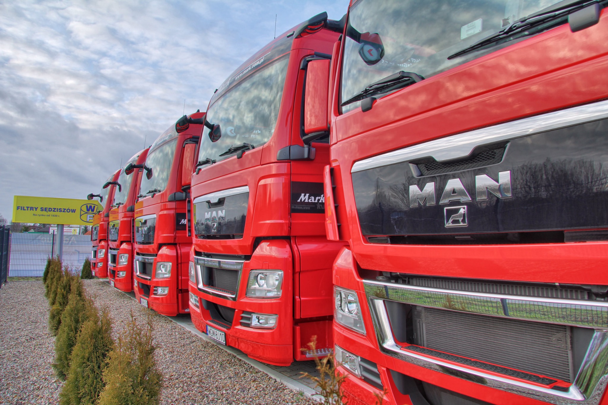 Flota czerwonych ciężarówek MAN ustawiona w rzędzie, widok z boku. Na pierwszym planie fragment reklamy filtrów. Pochmurne niebo w tle.
