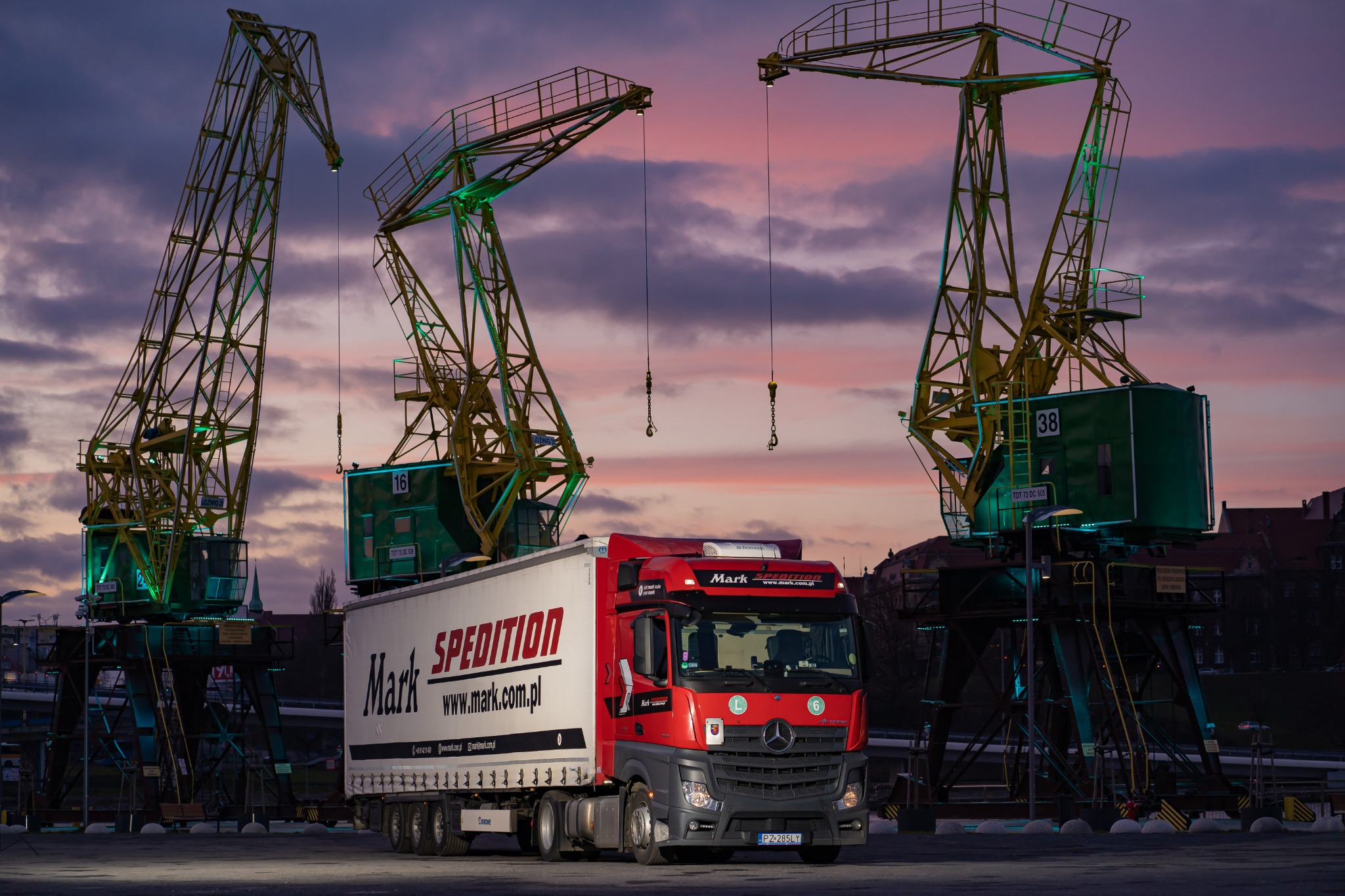 Ciężarówka Mark Spedition na tle portowych żurawi o zmierzchu. Czerwono-biała kabina z logo, naczepa z nazwą firmy. Widoczne detale: tablice rejestracyjne, oświetlenie.