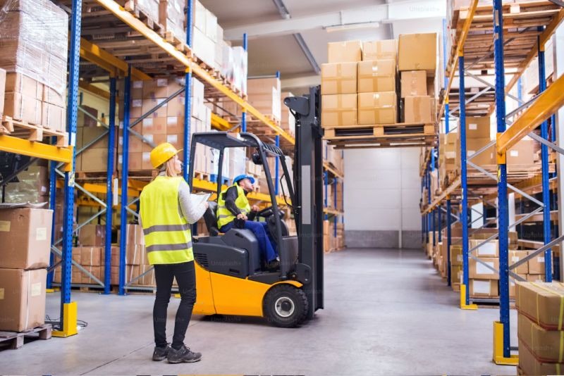 Wnętrze magazynu: operator wózka widłowego transportuje kartony na palecie, nadzorowany przez pracownika z notatnikiem. Wysokie regały wypełnione towarem.