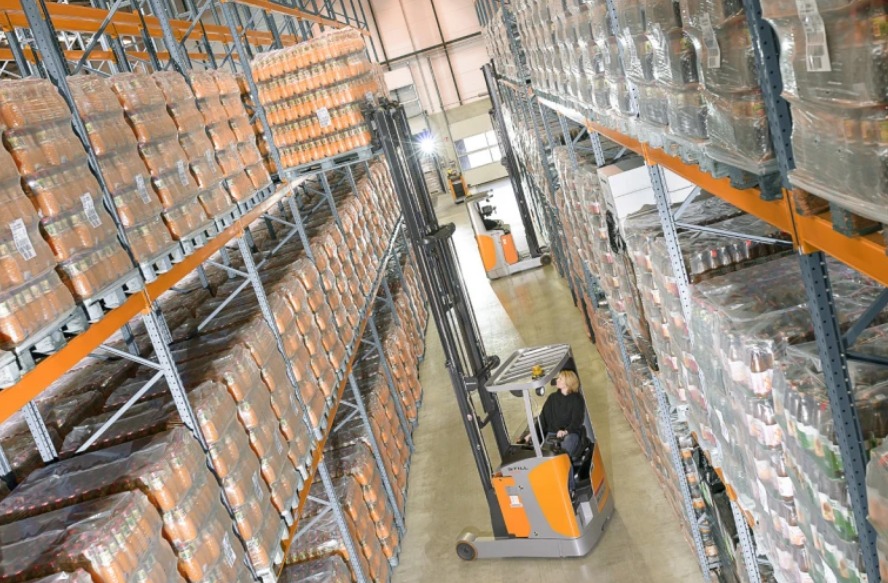 Wysoki magazyn z regałami wypełnionymi zapakowanymi produktami. Wąski wózek widłowy STILL z operatorem przemieszcza paletę z towarem. Długi kadr z perspektywą.