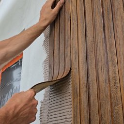 NOE - Montaż drewnopodobnych paneli elewacyjnych na ścianie z użyciem zaprawy. Widoczne dłonie wykonawcy i fragment okna zabezpieczonego folią.