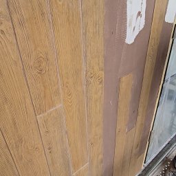 NOE - Montaż paneli elewacyjnych imitujących drewno na brązowej ścianie z fragmentami białej zaprawy. Widoczne narzędzia i fragment okna.