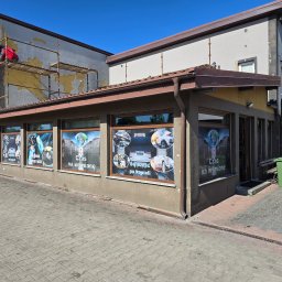 NOE - Budynek usługowy z częściowo odnowioną elewacją. Na rusztowaniu pracownik w czerwonej kurtce. Okna z reklamami myjni samochodowej.