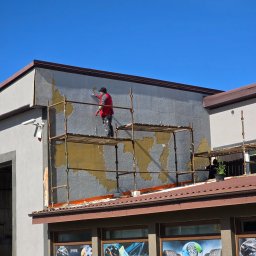 NOE - Pracownik w czerwonej kamizelce na rusztowaniu tynkuje szarą elewację budynku. Widoczne fragmenty starego tynku. W tle błękitne niebo.