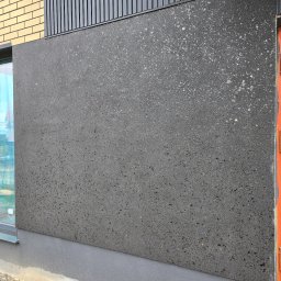 NOE - Szara elewacja z fakturą, połączona z elementami drewnianymi i ceglanymi. Widoczny fragment okna i podmurówka. Surowy, nowoczesny styl.