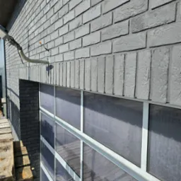 Szara elewacja z cegły klinkierowej, widoczna z bliska, z fragmentem rusztowania po lewej stronie. Okno z białymi ramami pod elewacją.