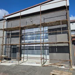 Szara ceglana elewacja budynku w trakcie prac, z widocznymi rusztowaniami i fragmentem pomalowanej ściany. Częściowo widoczne okna i niebieskie niebo w tle.