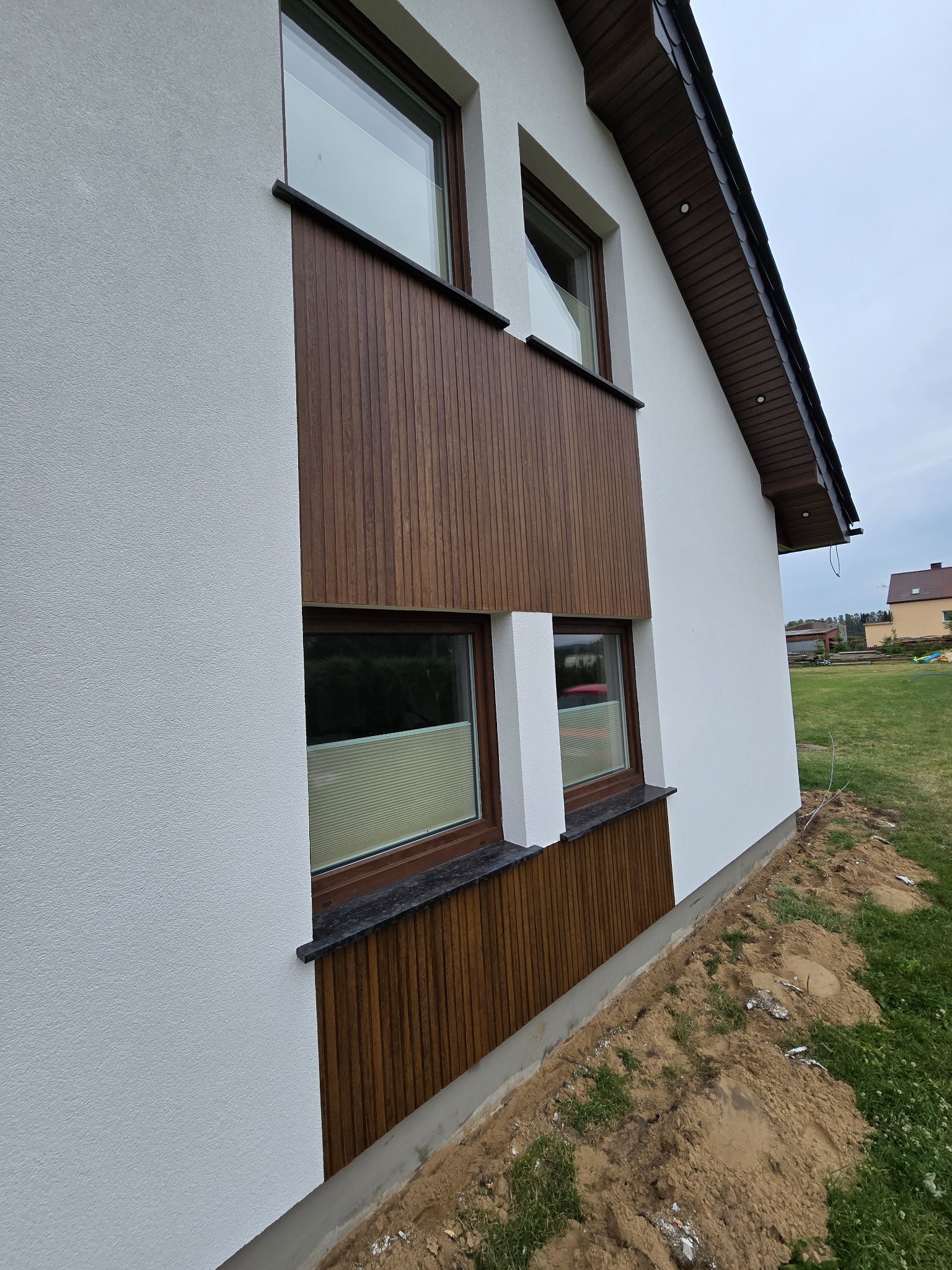 Boczny widok domu z białą elewacją i drewnianymi panelami pod oknami, brązowe okna, widoczne trawniki i sąsiedni dom w oddali, pochmurne niebo.