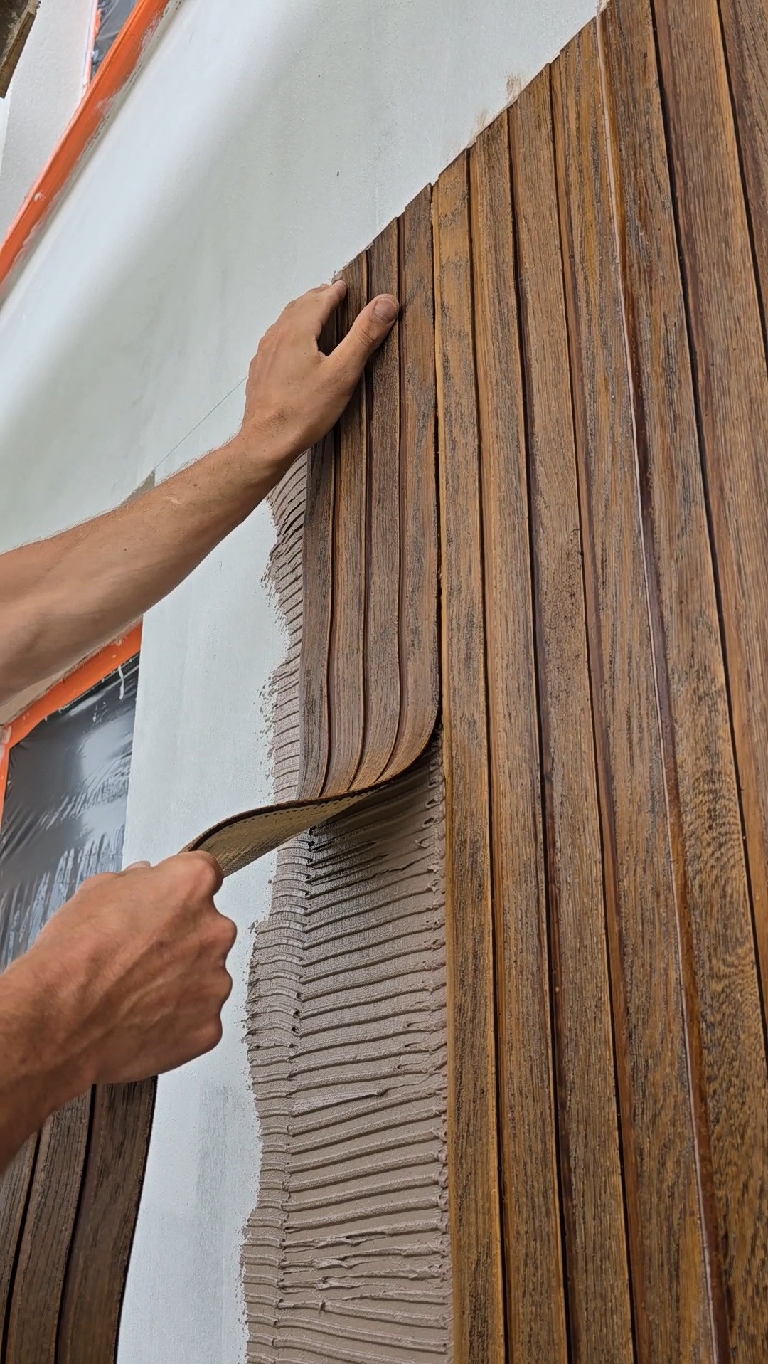 Montaż drewnopodobnych paneli elewacyjnych na ścianie z użyciem zaprawy. Widoczne dłonie wykonawcy i fragment okna zabezpieczonego folią.