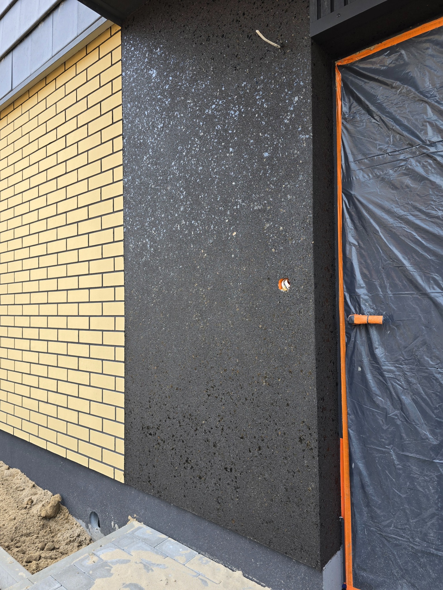 Fragment elewacji budynku z wykończeniem z żółtej cegły i ciemnoszarego tynku strukturalnego. Okno zabezpieczone folią malarską.