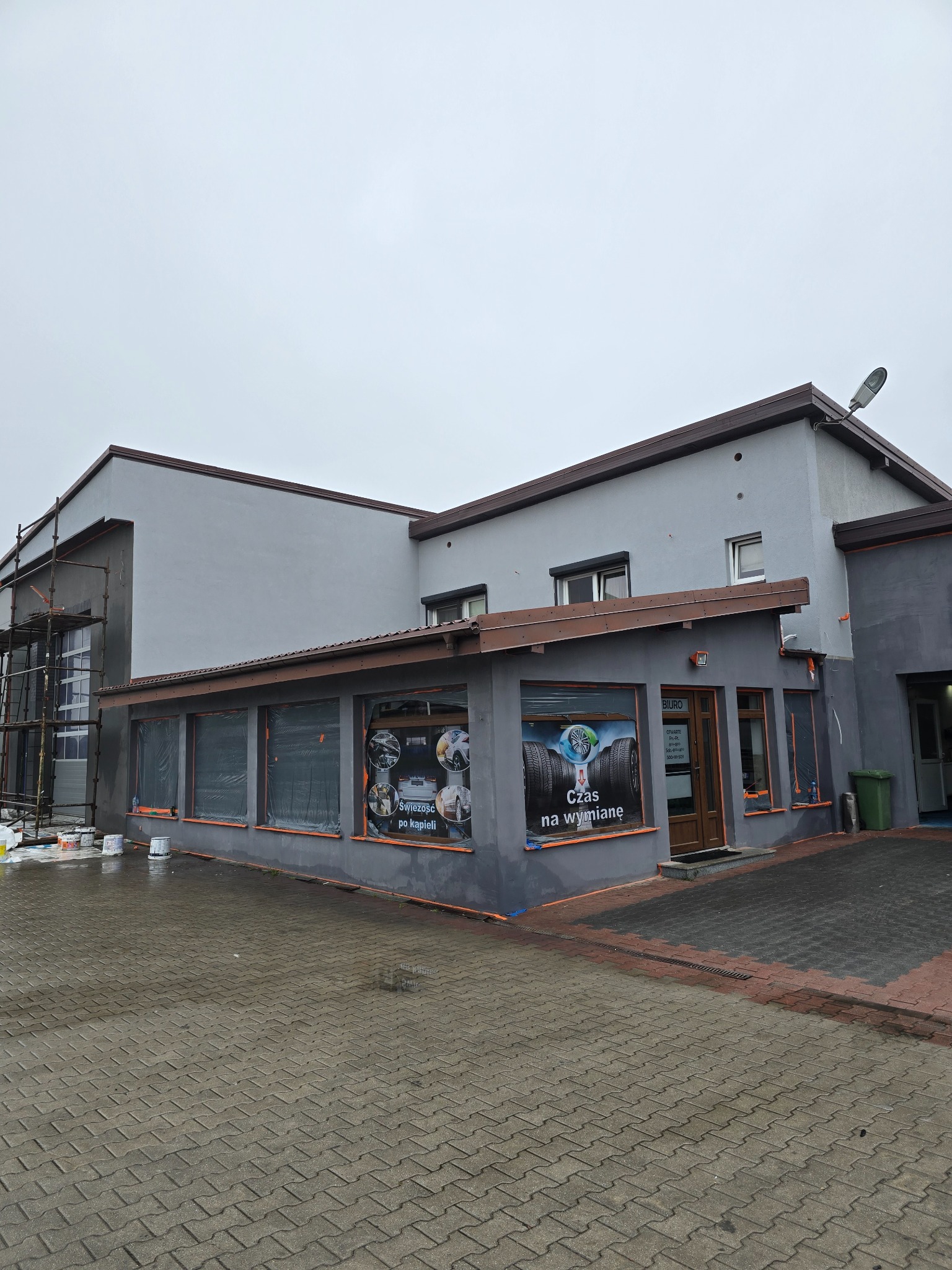 Szara elewacja budynku usługowego w trakcie malowania, widoczne rusztowania i puszki z farbą. Okna zakryte folią, na szybach reklamy opon.