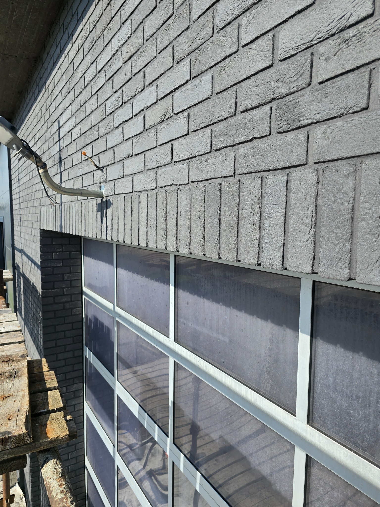 Szara elewacja z cegły klinkierowej, widoczna z bliska, z fragmentem rusztowania po lewej stronie. Okno z białymi ramami pod elewacją.