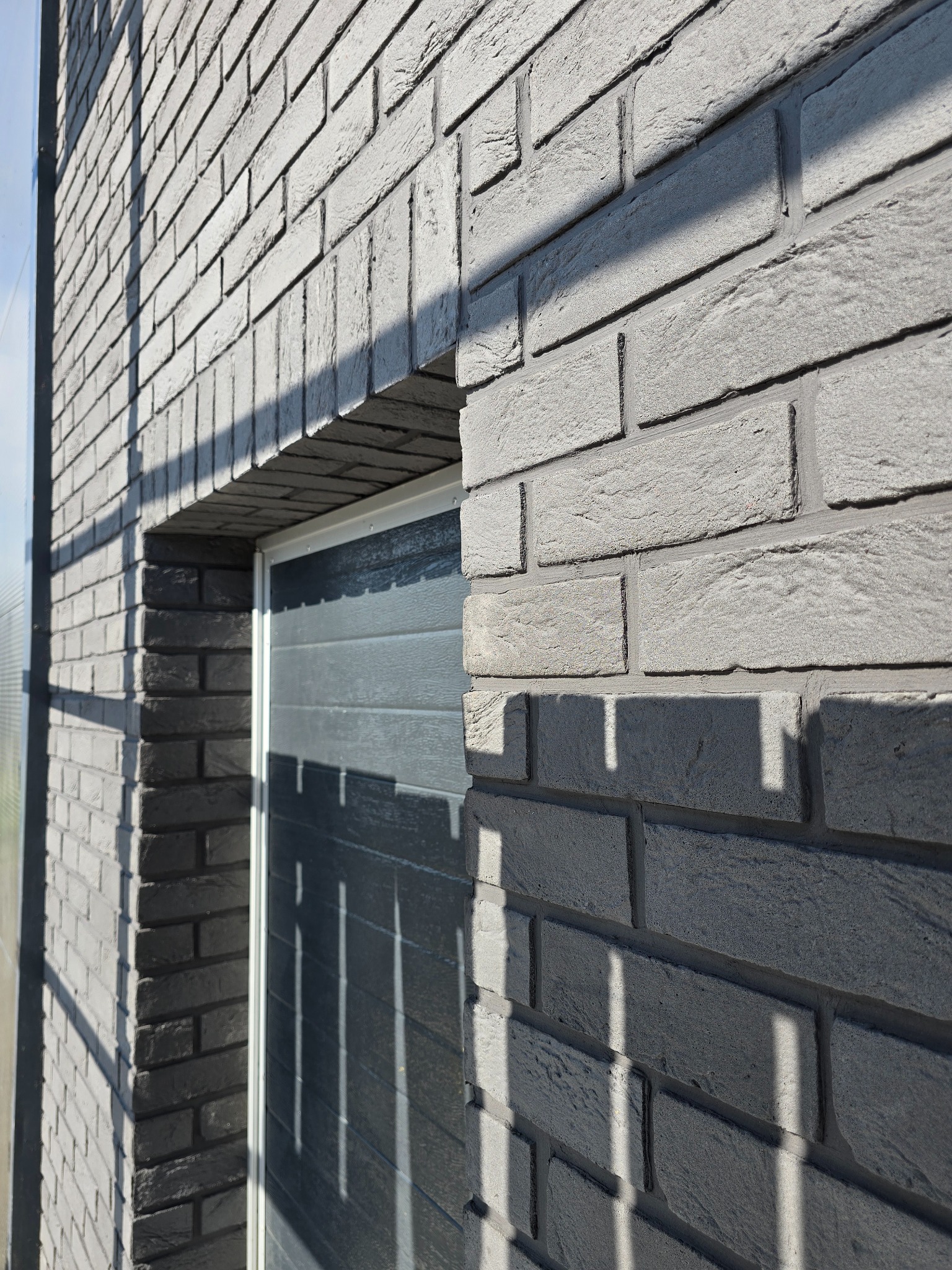 Szara elewacja z cegły klinkierowej wokół okna garażowego, oświetlona popołudniowym słońcem, z widoczną fakturą i cieniowaniem.