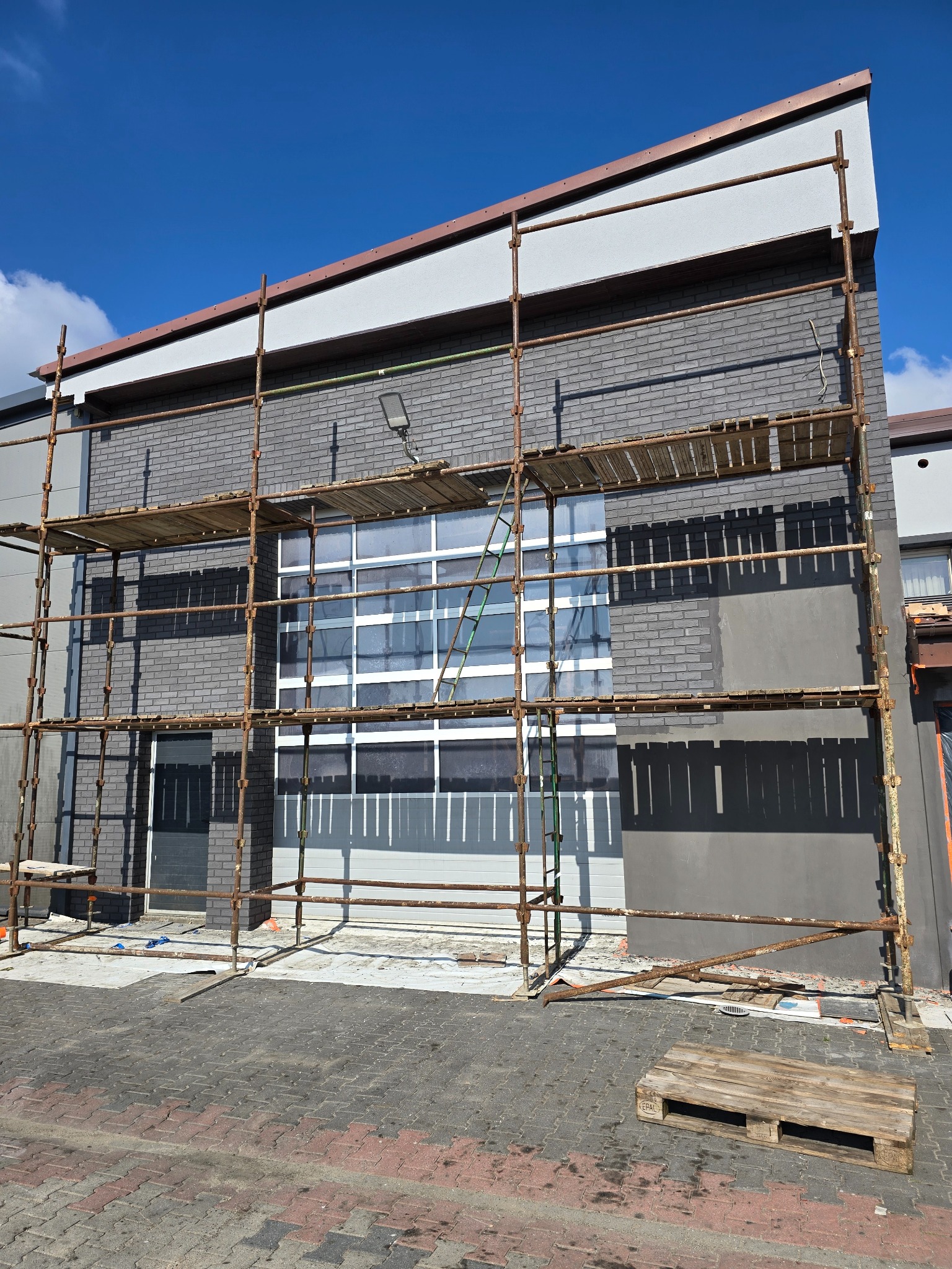Szara ceglana elewacja budynku w trakcie prac, z widocznymi rusztowaniami i fragmentem pomalowanej ściany. Częściowo widoczne okna i niebieskie niebo w tle.