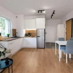 Jasna kuchnia z białymi szafkami i drewnianym blatem, uzupełniona o stół jadalny i szarą sofę, tworząca spójną przestrzeń dzienną w nowoczesnym stylu.