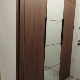 Nowoczesna szafa wnękowa z drewnianymi frontami i lustrzanymi panelami. Minimalistyczny design, czarne uchwyty, jasna ściana i marmurowa podłoga w tle.