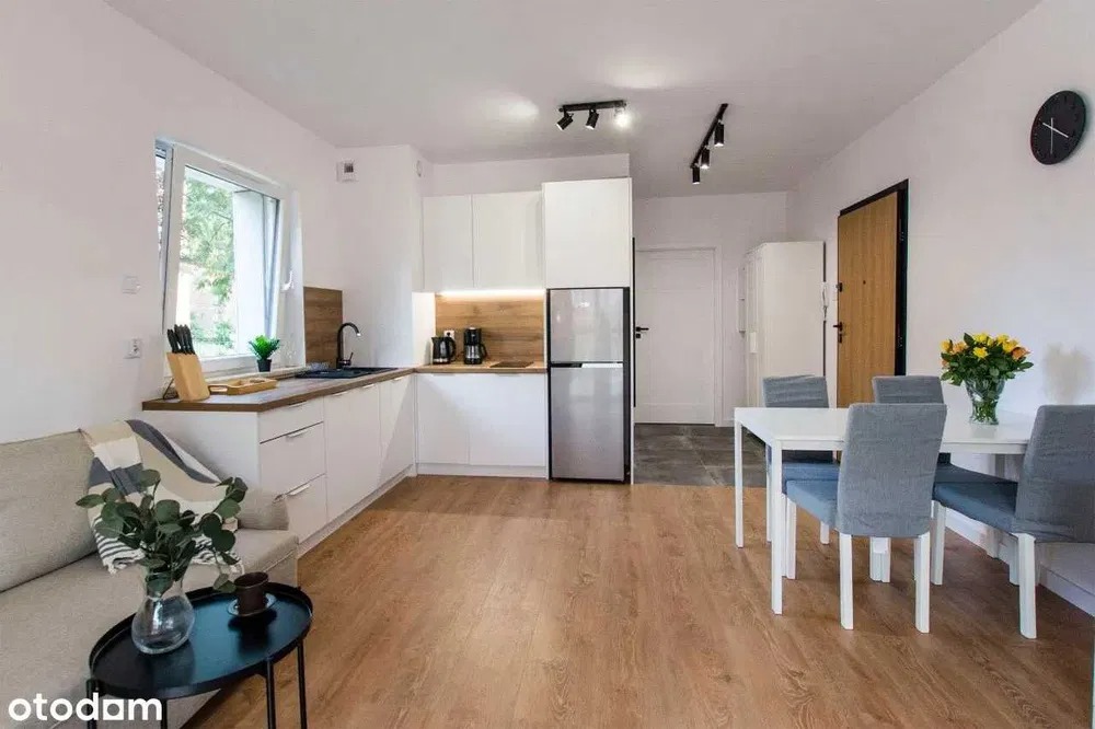 Jasna kuchnia z białymi szafkami i drewnianym blatem, uzupełniona o stół jadalny i szarą sofę, tworząca spójną przestrzeń dzienną w nowoczesnym stylu.