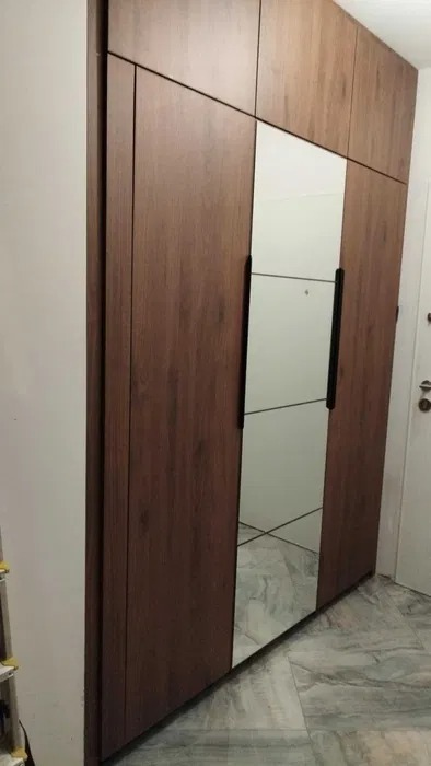 Nowoczesna szafa wnękowa z drewnianymi frontami i lustrzanymi panelami. Minimalistyczny design, czarne uchwyty, jasna ściana i marmurowa podłoga w tle.