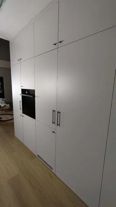 Biała szafa wnękowa z czarnym piekarnikiem i uchwytami, wbudowana w ścianę, widok z dołu. Minimalistyczny design, jasne drewniane panele podłogowe.