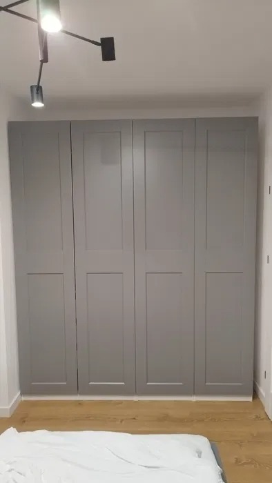 Szara, czterodrzwiowa szafa wnękowa z panelowymi frontami, oświetlona nowoczesną lampą sufitową, na tle drewnianej podłogi i białej pościeli.