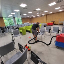 DOREK Yury Volkau - Pracownik w czarnej koszulce czyści szarą tapicerkę krzesła w sali konferencyjnej za pomocą odkurzacza piorącego; widoczne dodatkowe krzesła, sofy i sprzęt czyszczący.
