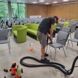 DOREK Yury Volkau - Mężczyzna w czarnej koszulce i szortach czyści tapicerkę krzesła w sali konferencyjnej za pomocą elektrycznej szczotki, widoczne butelki z detergentami, spryskiwacze i odkurzacz piorący.