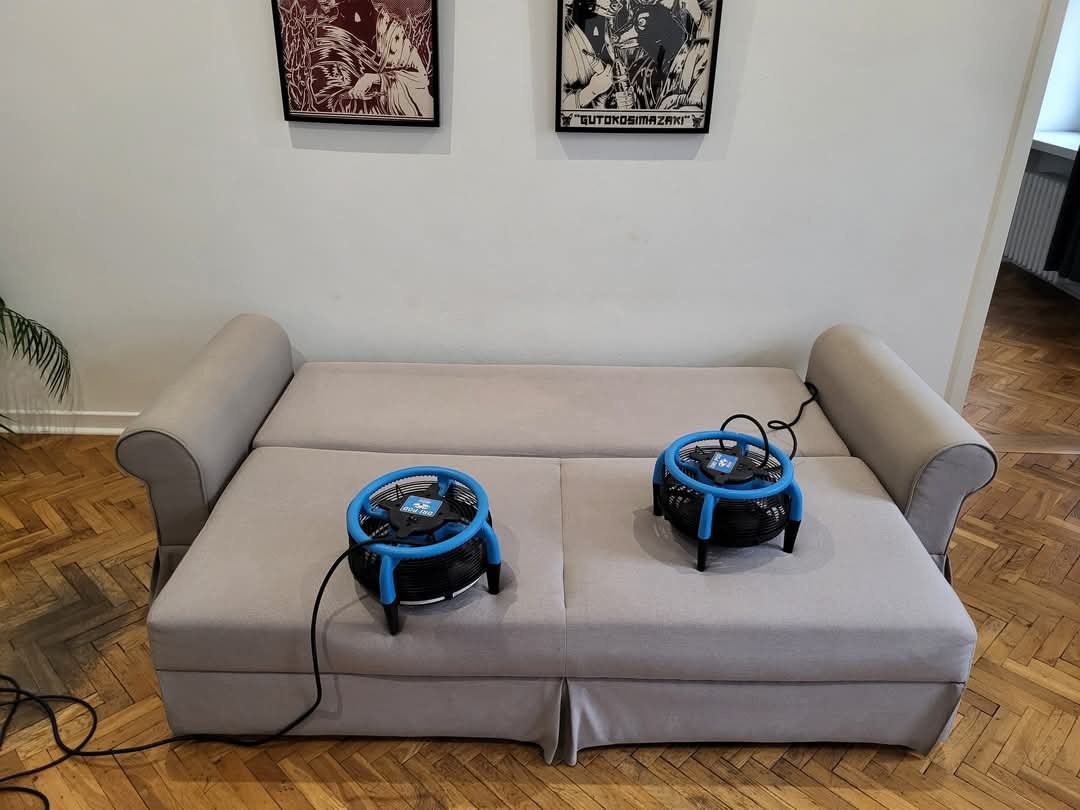 Jasnoszara rozkładana kanapa z dwoma niebieskimi wentylatorami do suszenia po praniu, wiszące grafiki na ścianie w tle.