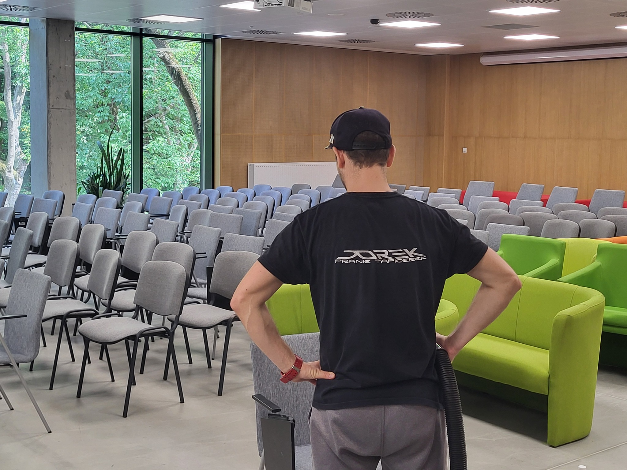 Pracownik firmy czyszczącej, z odkurzaczem w dłoni, stoi w sali konferencyjnej z rzędami krzeseł i zieloną kanapą, prawdopodobnie przed rozpoczęciem lub po zakończeniu czyszczenia tapicerki.
