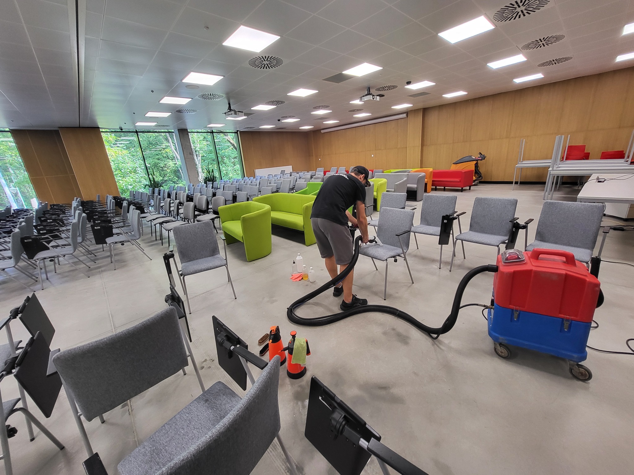 Pracownik w czarnej koszulce czyści szarą tapicerkę krzesła w sali konferencyjnej za pomocą odkurzacza piorącego; widoczne dodatkowe krzesła, sofy i sprzęt czyszczący.