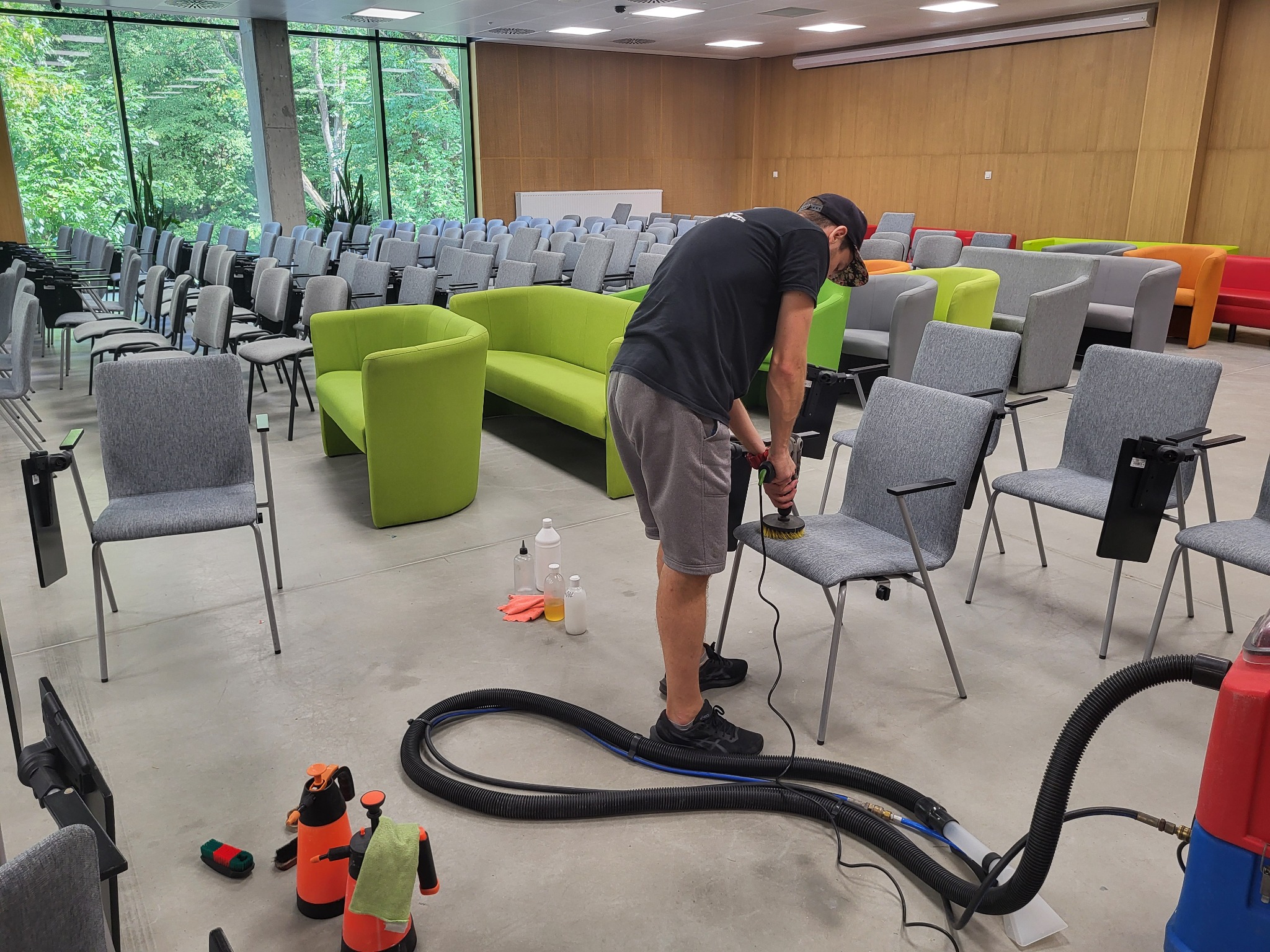 Mężczyzna w czarnej koszulce i szortach czyści tapicerkę krzesła w sali konferencyjnej za pomocą elektrycznej szczotki, widoczne butelki z detergentami, spryskiwacze i odkurzacz piorący.